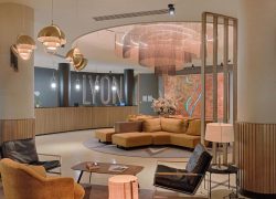 Lobby d'hôtel avec comptoir de réception en bois adossé à une enseigne lumineuse « Lyon » au milieu, canapé incurvé jaune moutarde autour d'une table basse sur tapis circulaire, fauteuils en cuir noir et orange au premier plan, suspensions en dôme doré et grand lustre à étages au-dessus, et œuvre en forme de carte sur un écran à claire-voie à droite, un lobby accueillant pour un hôtel pour séminaire.