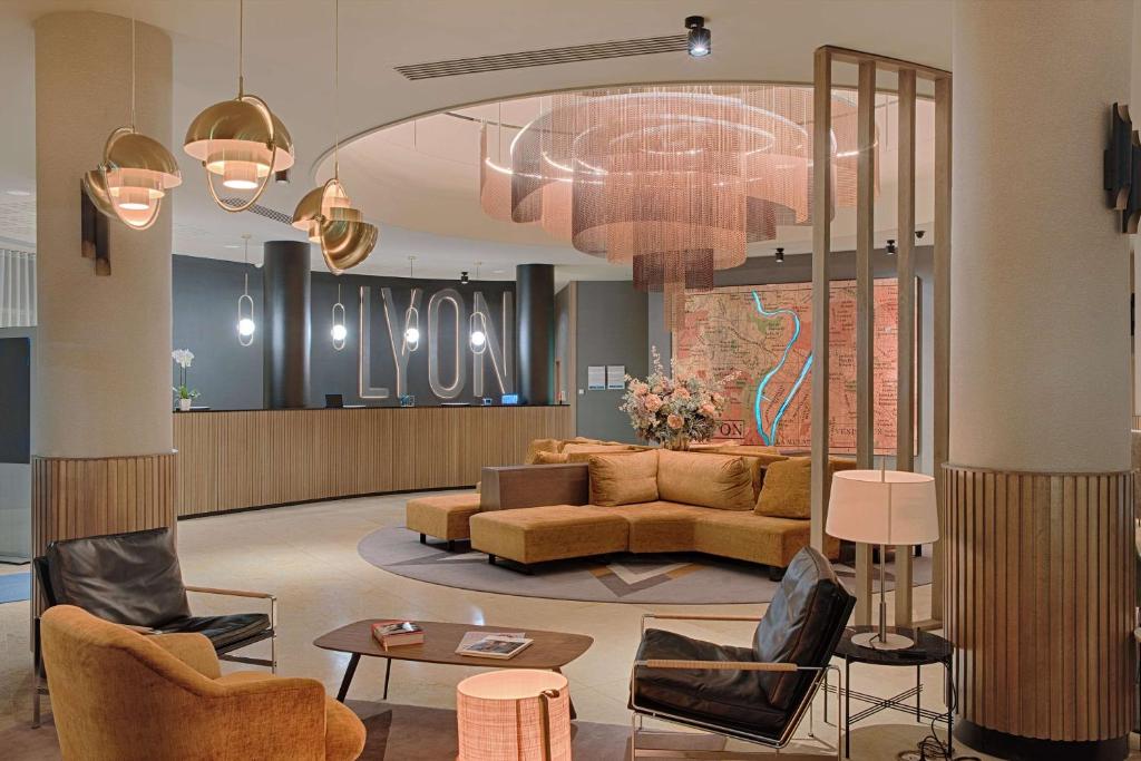 Lobby d'hôtel avec comptoir de réception en bois adossé à une enseigne lumineuse « Lyon » au milieu, canapé incurvé jaune moutarde autour d'une table basse sur tapis circulaire, fauteuils en cuir noir et orange au premier plan, suspensions en dôme doré et grand lustre à étages au-dessus, et œuvre en forme de carte sur un écran à claire-voie à droite, un lobby accueillant pour un hôtel pour séminaire.