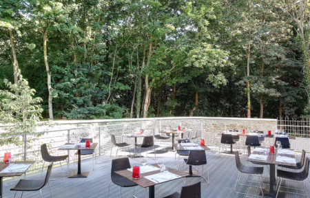 Outdoor wooden terrace ideal for a green seminar, with wooden tables set with white napkins, glasses and red candle holders and dark woven chairs in the foreground, a stone balustrade with metal railing along the edge, and a dense backdrop of tall green trees under daylight.