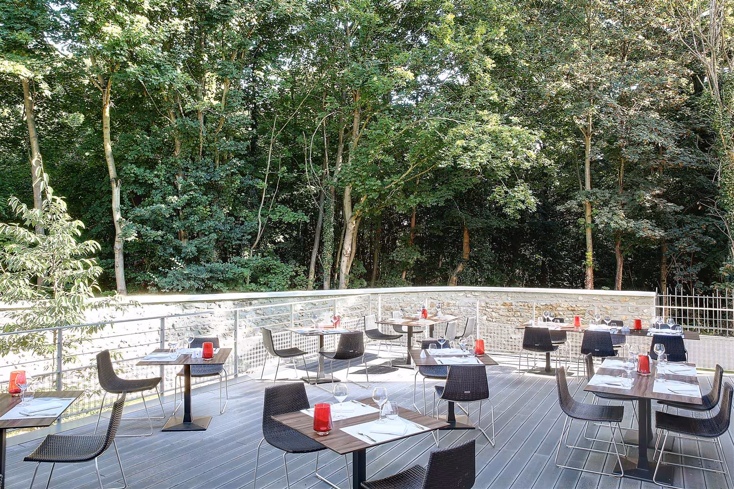 Outdoor wooden terrace ideal for a green seminar, with wooden tables set with white napkins, glasses and red candle holders and dark woven chairs in the foreground, a stone balustrade with metal railing along the edge, and a dense backdrop of tall green trees under daylight.