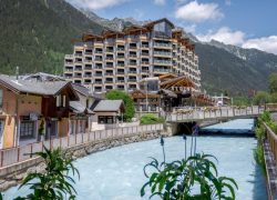 en::Corner hotel building with nine rows of rooms and balconies on each side, overlooking the Arve River, its bridge and the mountains. fr::Bâtiment d’hôtel en angle avec neuf rangées de chambres et balcons de chaque côté, vue sur l’Arve, son pont et les montagnes.