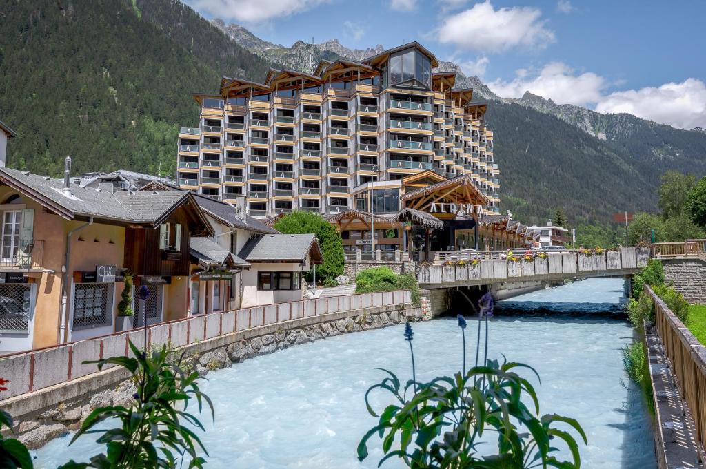 en::Corner hotel building with nine rows of rooms and balconies on each side, overlooking the Arve River, its bridge and the mountains. fr::Bâtiment d’hôtel en angle avec neuf rangées de chambres et balcons de chaque côté, vue sur l’Arve, son pont et les montagnes.