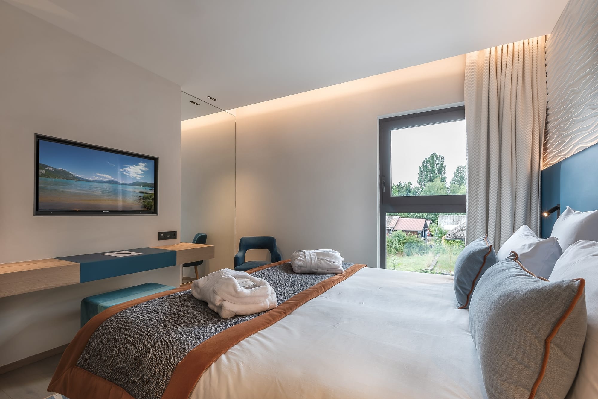 Hotel guest room for group accommodation with a double bed and folded bathrobes in the foreground, a light wood desk with blue storage and a wall-mounted television on the left, a blue armchair in the background, and a large window with white curtains overlooking trees on the right.