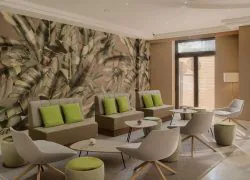 Salon d'hôtel avec grande fresque murale à motif de feuilles tropicales à gauche, canapés beiges aux coussins verts et fauteuils blancs incurvés disposés autour de petites tables basses rondes et de poufs rembourrés verts au premier plan, porte vitrée donnant sur l'extérieur et lampadaire à droite, un salon relaxant adapté à un séminaire au vert.