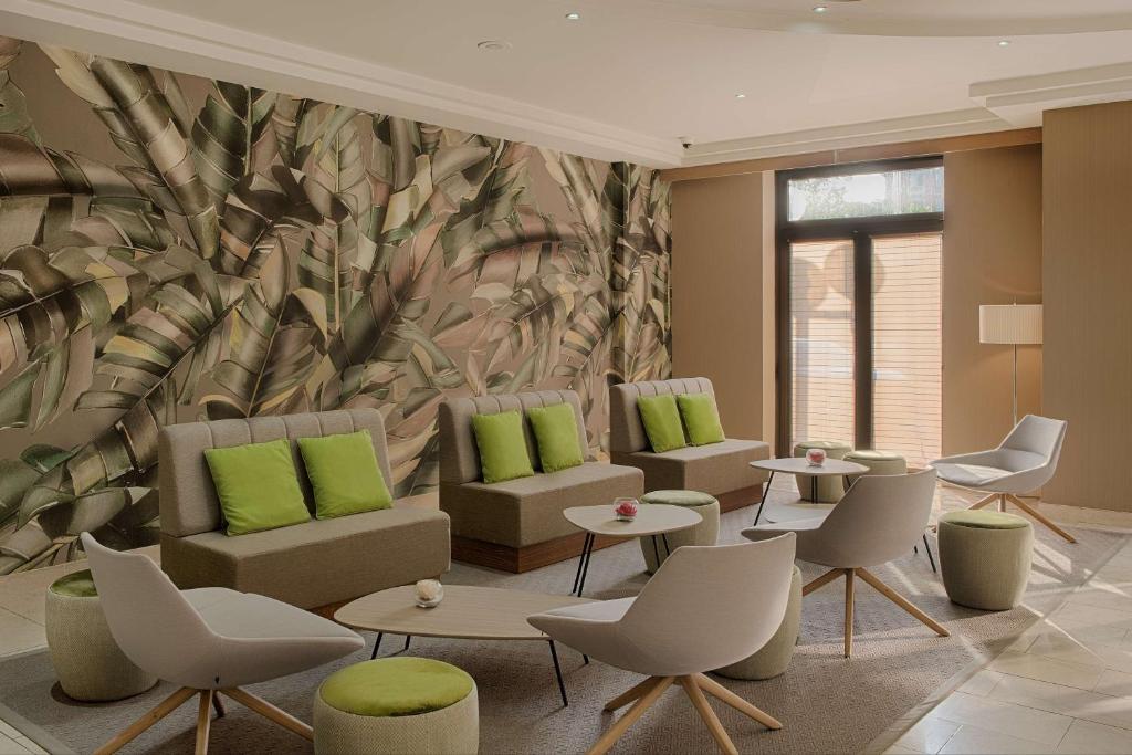 Hotel lounge with a large botanical leaf-patterned wall mural on the left, beige sofas with green cushions and white curved armchairs arranged around small round coffee tables and green upholstered poufs in the foreground, a glass door leading outside and a floor lamp on the right, a relaxing lounge suited for a green seminar.