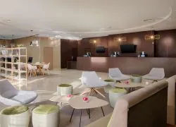 Lobby-lounge d'hôtel avec fauteuils gris et petites tables basses en bois entourées de poufs rembourrés verts au premier plan, comptoir de réception en bois sombre avec écrans et suspensions géométriques en arrière, et étagère ouverte avec plantes près d'un petit coin repas à gauche, un lobby accueillant pour un hôtel pour séminaire.