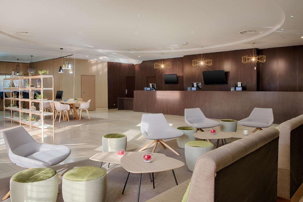 Hotel lobby lounge with grey armchairs and small wooden coffee tables surrounded by green upholstered poufs in the foreground, a dark wood-panelled reception desk with screens and geometric pendant lights behind, and an open shelving unit with plants beside a small dining area on the left, a welcoming lobby for a seminar hotel.
