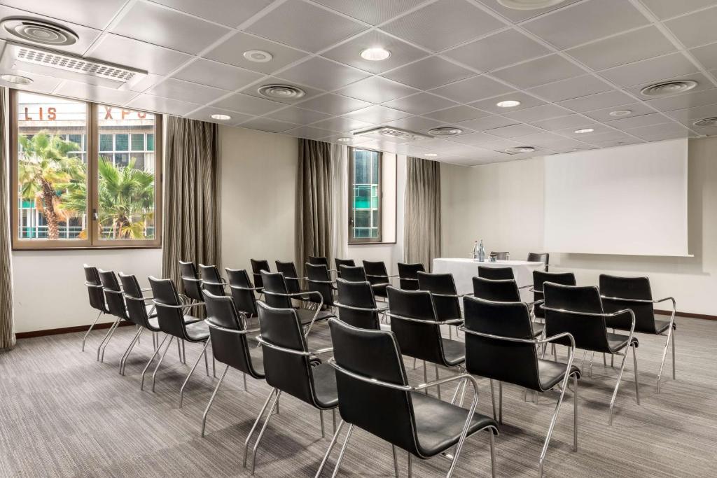 Meeting room with rows of black leather chairs with chrome frames facing a projection screen on the right, a white speaker's table with glasses and a carafe at the front, beige curtains framing windows with palm trees visible on the left, and a grey checkered ceiling with recessed lighting above, a room configured for room rental.