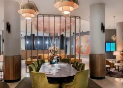 Hotel lobby with an oval dark wooden table surrounded by olive green velvet chairs in the foreground, brass dome pendant lights hanging above, fluted wooden-clad columns on either side, a decorative chandelier suspended within a slatted brass screen, and an orange sofa with a large map artwork on the wall in the background, a welcoming lobby for a seminar hotel.