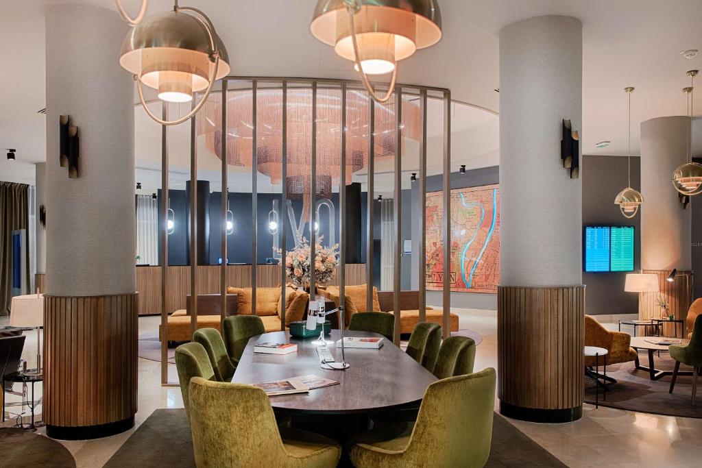 Hotel lobby with an oval dark wooden table surrounded by olive green velvet chairs in the foreground, brass dome pendant lights hanging above, fluted wooden-clad columns on either side, a decorative chandelier suspended within a slatted brass screen, and an orange sofa with a large map artwork on the wall in the background, a welcoming lobby for a seminar hotel.