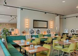 Restaurant d'hôtel avec banquette verte et chaises rembourrées jaune-vert autour de tables en bois dressées de couverts et bougies au premier plan, photographies rondes encadrées en noir et blanc et miroir rectangulaire entre des appliques murales au-dessus, et plantes en pot hautes à gauche, un restaurant proposant une restauration sur place.