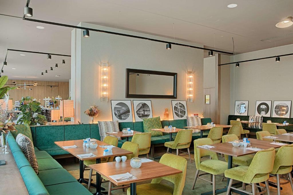 Restaurant d'hôtel avec banquette verte et chaises rembourrées jaune-vert autour de tables en bois dressées de couverts et bougies au premier plan, photographies rondes encadrées en noir et blanc et miroir rectangulaire entre des appliques murales au-dessus, et plantes en pot hautes à gauche, un restaurant proposant une restauration sur place.