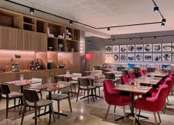 Restaurant d'hôtel avec fauteuils en velours rouge et chaises grises autour de tables en bois dressées de verrerie et bougies au premier plan, banquette à motifs avec coussins colorés le long du mur, photographies rondes encadrées en noir et blanc au-dessus, et étagères en bois ouvertes présentant bouteilles et objets décoratifs à gauche, un restaurant proposant une restauration sur place.