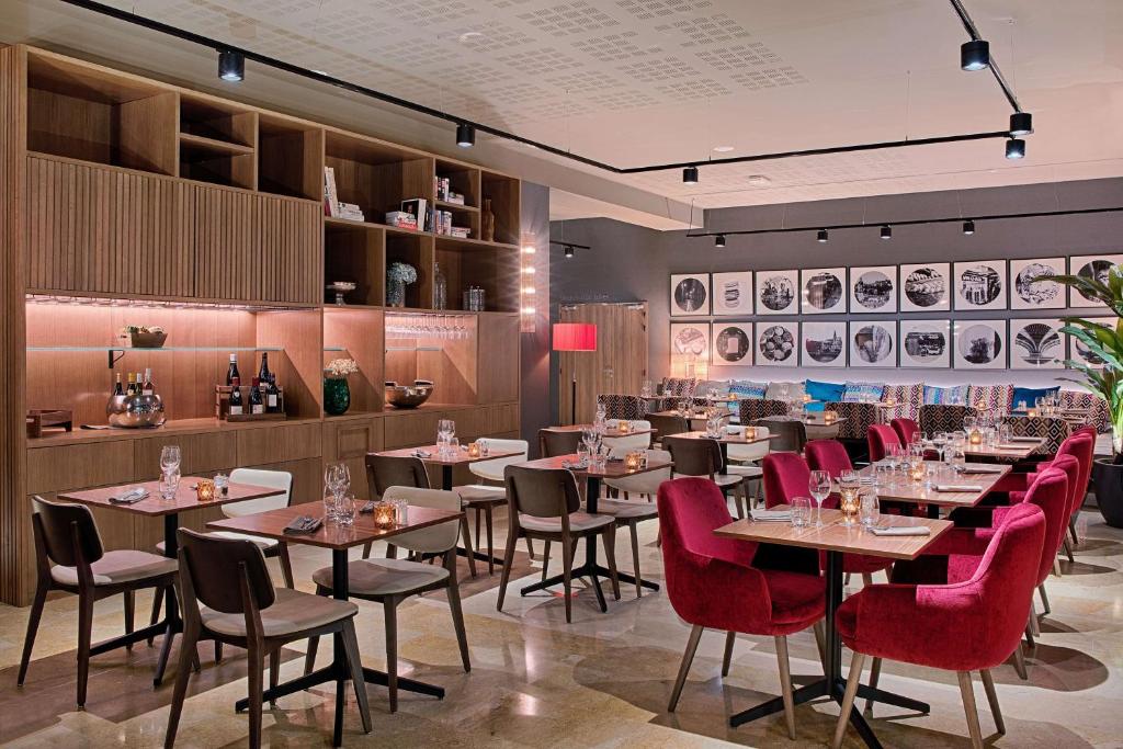 Restaurant d'hôtel avec fauteuils en velours rouge et chaises grises autour de tables en bois dressées de verrerie et bougies au premier plan, banquette à motifs avec coussins colorés le long du mur, photographies rondes encadrées en noir et blanc au-dessus, et étagères en bois ouvertes présentant bouteilles et objets décoratifs à gauche, un restaurant proposant une restauration sur place.