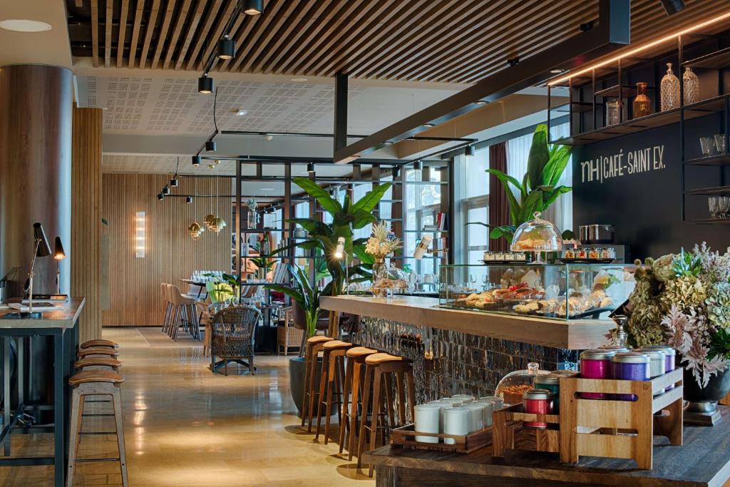 Hotel café with a stone-clad counter displaying pastries and sandwiches under glass domes in the foreground, wooden bar stools lined up along the counter, a wall sign reading "NH Café-Saint EX." on the right, tall potted plants and hanging pendant lights above, and open wooden shelving with glassware and decorative bottles in the background, a café offering on-site dining.