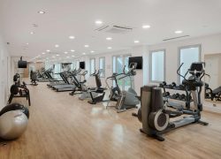 Salle de sport d'hôtel avec rangées d'appareils cardio dont tapis de course, vélos elliptiques et vélos d'appartement chacun équipé d'un petit écran, alignés sur un parquet au premier plan, ballons de gymnastique et porte-poids à gauche, et grand miroir et porte-haltères à droite, une salle de fitness adaptée à un séminaire bien-être.