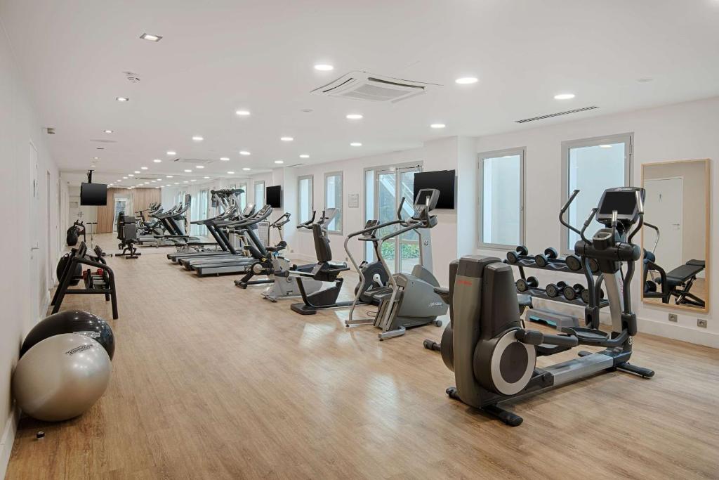 Salle de sport d'hôtel avec rangées d'appareils cardio dont tapis de course, vélos elliptiques et vélos d'appartement chacun équipé d'un petit écran, alignés sur un parquet au premier plan, ballons de gymnastique et porte-poids à gauche, et grand miroir et porte-haltères à droite, une salle de fitness adaptée à un séminaire bien-être.