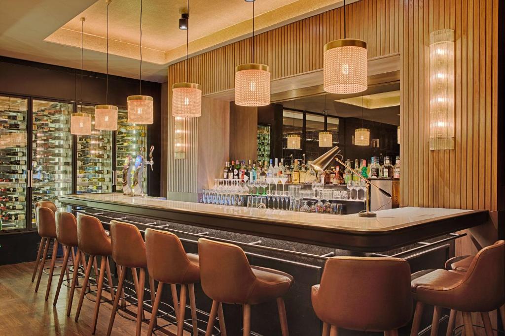 Hotel bar with brown leather high stools lining a curved black counter topped with marble in the foreground, shelves of bottles and glassware behind, glass-doored wine refrigerators on the left, and woven cylindrical pendant lights hanging above, a bar area suited for an afterwork.