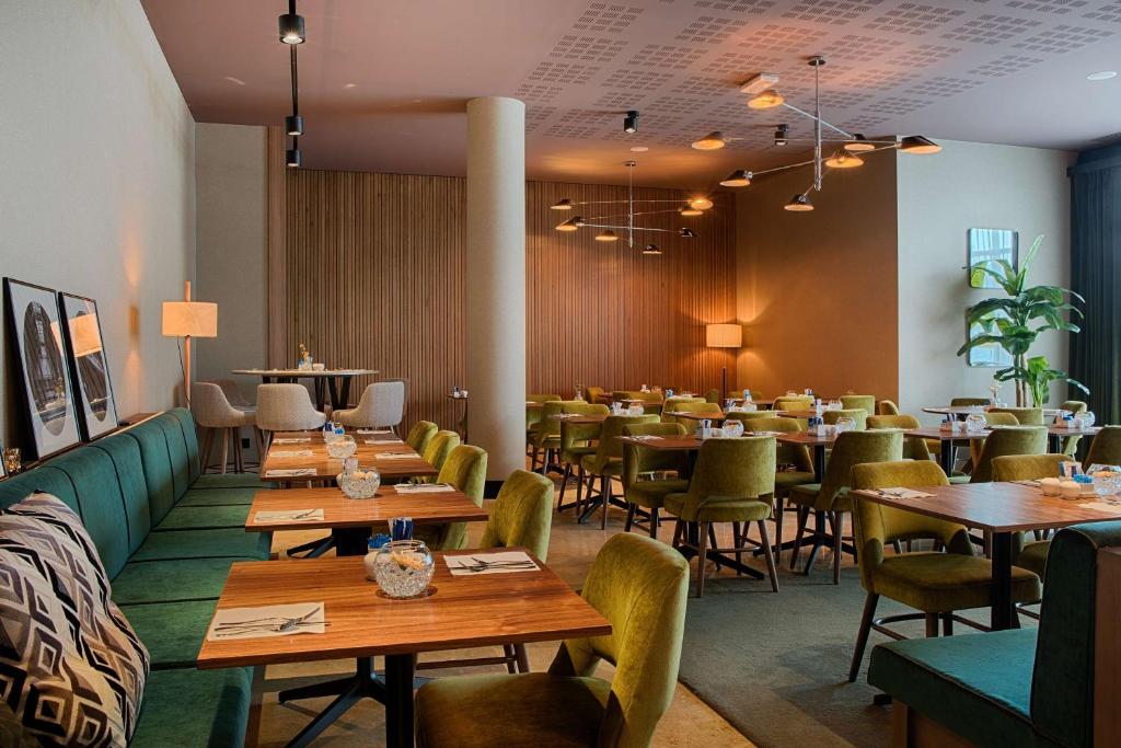Hotel restaurant with a green banquette and olive velvet chairs around wooden tables set with cutlery and glass candle holders in the foreground, wooden panelled and terracotta-toned walls in the middle ground, a sculptural multi-arm pendant light fixture hanging above, and framed black-and-white photographs on the left wall, a restaurant offering on-site dining.