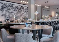 Hotel restaurant with a round wooden table set with cutlery, napkins and a small vase in the foreground, grey herringbone-patterned chairs surrounding it, a long buffet counter with high stools and a black-and-white forest mural on the wall in the middle ground, and spotlights and wall sconces above, a restaurant offering on-site dining.