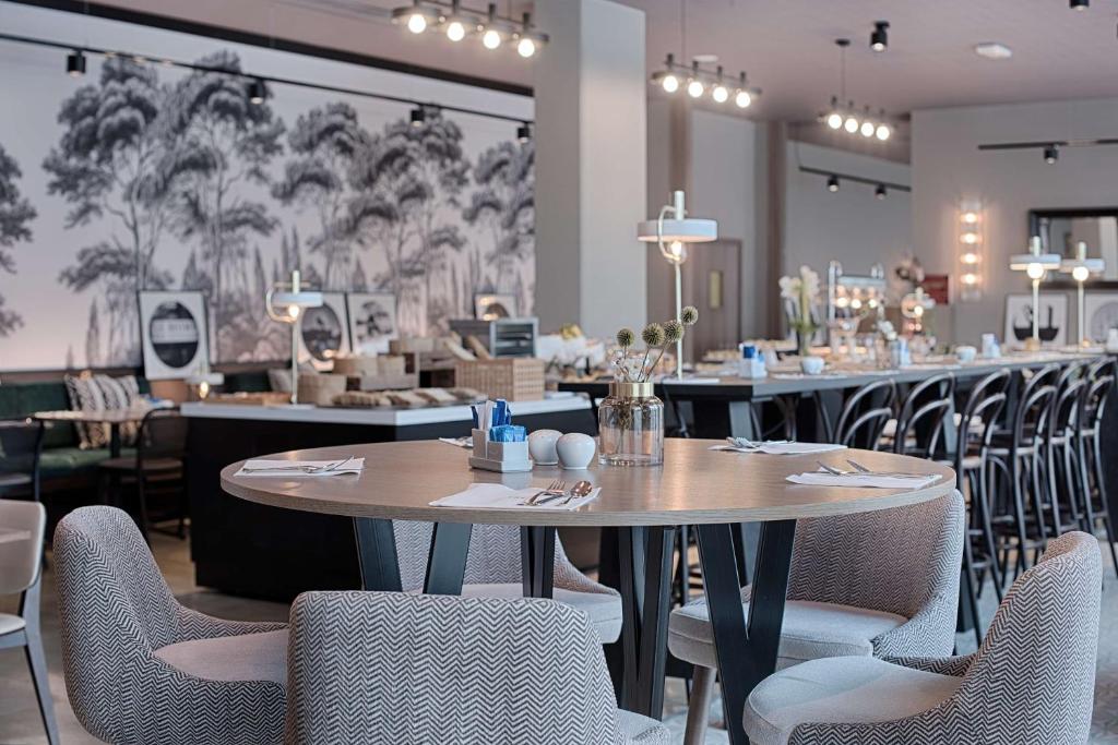 Hotel restaurant with a round wooden table set with cutlery, napkins and a small vase in the foreground, grey herringbone-patterned chairs surrounding it, a long buffet counter with high stools and a black-and-white forest mural on the wall in the middle ground, and spotlights and wall sconces above, a restaurant offering on-site dining.