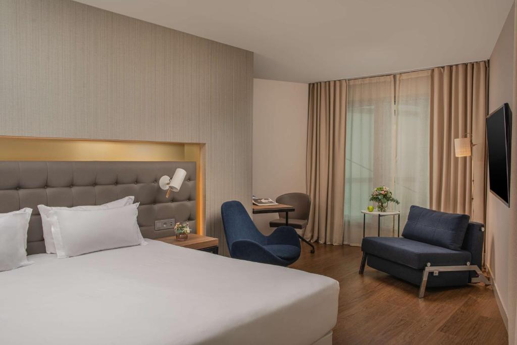 Chambre d'hôtel avec lit aux draps blancs et tête de lit rembourrée grise équipée d'une liseuse murale au premier plan, fauteuil rembourré bleu près d'un bureau en bois avec chaise pivotante au milieu, et canapé-lit bleu près d'une table d'appoint ronde avec fleurs et télévision murale près de fenêtres rideautées en arrière-plan, une chambre adaptée à un hôtel pour séminaire.