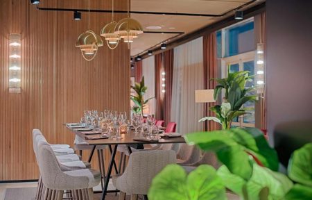 Restaurant d'hôtel avec longue table en bois sombre dressée de verres à vin et serviettes pliées au premier plan, chaises rembourrées grises et chaises roses de part et d'autre, suspensions en dôme de laiton au-dessus, murs en panneaux de bois avec bandeaux lumineux verticaux à gauche, et grandes plantes en pot et rideaux voilés en arrière-plan, un restaurant proposant une restauration sur place.