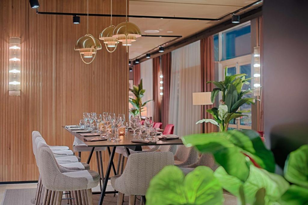 Hotel restaurant with a long dark wooden table set with wine glasses and folded napkins in the foreground, grey upholstered chairs and pink chairs on either side, brass dome pendant lights hanging above, wooden panelled walls with vertical lighting strips on the left, and large potted plants and sheer curtains in the background, a restaurant offering on-site dining.