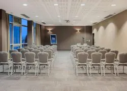 Salle de réunion avec rangées de chaises rembourrées grises aux pieds chromés séparées par une allée centrale face à une enseigne « NH Hotels » à l'enseigne de l'hôtel et petite table d'intervenant à l'avant, appliques murales et baies vitrées du sol au plafond à gauche, et plafond à caissons gris avec spots encastrés au-dessus, une salle configurée pour une location de salle.