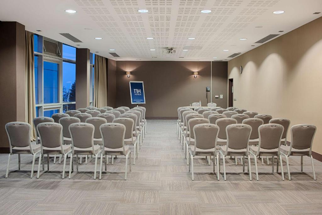 Salle de réunion avec rangées de chaises rembourrées grises aux pieds chromés séparées par une allée centrale face à une enseigne « NH Hotels » à l'enseigne de l'hôtel et petite table d'intervenant à l'avant, appliques murales et baies vitrées du sol au plafond à gauche, et plafond à caissons gris avec spots encastrés au-dessus, une salle configurée pour une location de salle.