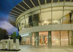 Vue extérieure nocturne d'un bâtiment d'hôtel vitré incurvé avec enseigne lumineuse « Lyon Airport » au-dessus d'une entrée éclairée en rez-de-chaussée au premier plan, grande installation sculpturale en forme de lettres « NH » sur le parvis pavé à gauche, auvent en lattes en surplomb au-dessus, et espace repas éclairé visible à l'étage en arrière-plan, un hôtel pour séminaire proche de l'aéroport.