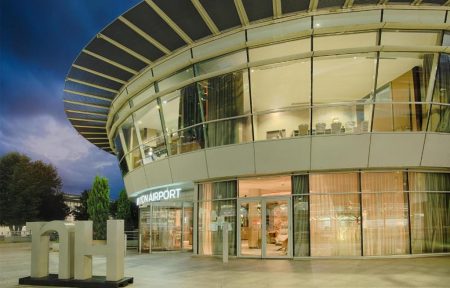 Night-time exterior view of a curved glass hotel building with an illuminated "Lyon Airport" sign above a lit ground-floor entrance in the foreground, a large sculptural "NH" letter installation on the paved forecourt on the left, an overhanging slatted canopy roof above, and a lit dining area visible on the upper floor in the background, a seminar hotel near the airport.