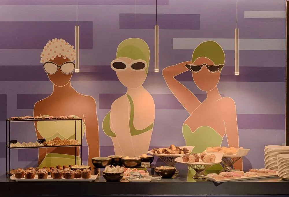 Espace buffet d'hôtel pour restauration sur place, avec des plateaux de viennoiseries et des bols sur un comptoir au premier plan, une grande fresque représentant trois nageuses stylisées sur un mur bleu à motifs géométriques en arrière-plan, et des suspensions cylindriques au-dessus.