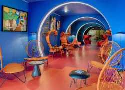 Couloir lounge d'hôtel dans l'esprit afterwork, comprenant des fauteuils en osier orange et rotin avec petites tables rondes noires sur un sol rouge au premier plan, un tunnel d'arches répétées en bleu avec tableaux pop-art encadrés au centre, et des assises rouges visibles au fond en arrière-plan.