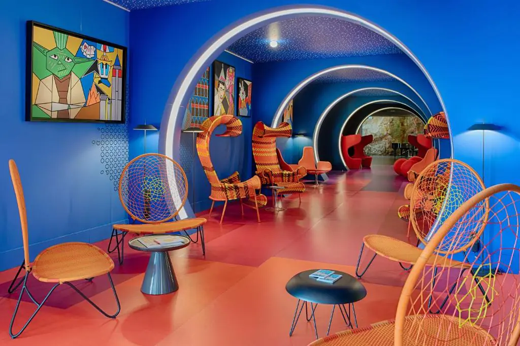 Couloir lounge d'hôtel dans l'esprit afterwork, comprenant des fauteuils en osier orange et rotin avec petites tables rondes noires sur un sol rouge au premier plan, un tunnel d'arches répétées en bleu avec tableaux pop-art encadrés au centre, et des assises rouges visibles au fond en arrière-plan.
