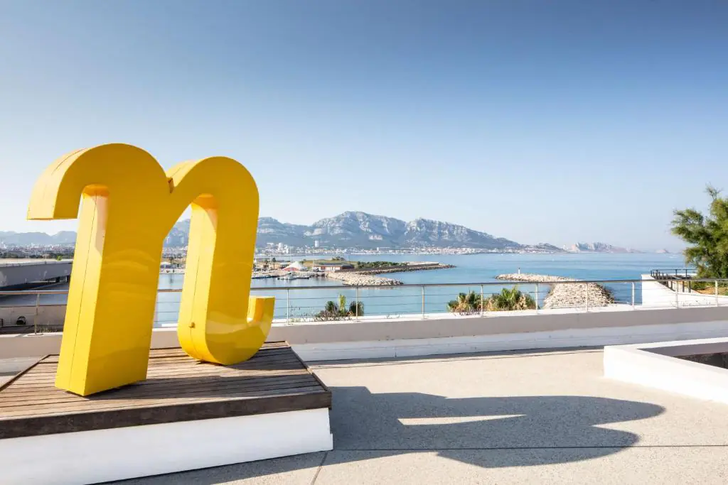Terrasse en rooftop incarnant l'esprit séminaire en bord de mer, comprenant une grande sculpture jaune en forme de « n » sur une plateforme en bois au premier plan, une rambarde en verre et métal au centre, et une baie côtière avec montagnes et port de plaisance sous un ciel dégagé en arrière-plan.