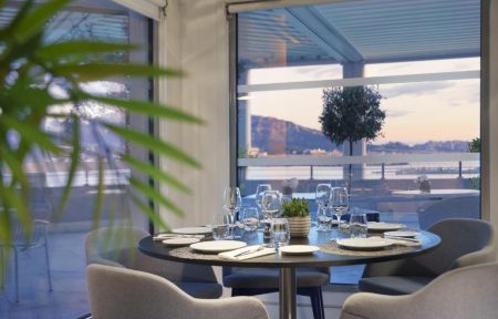 Restaurant d'hôtel pour restauration sur place avec une table ronde noire dressée de vaisselle blanche, verres et couverts au centre, des fauteuils gris rembourrés tout autour, de grandes baies vitrées donnant sur une terrasse et la mer à l'arrière-plan, et une plante verte au premier plan à gauche.