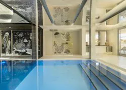 Piscine intérieure proposant une ambiance séminaire bien-être, comprenant un bassin rectangulaire bleu vif avec marches carrelées au premier plan, des fresques en mosaïque à motifs marins de méduses et pieuvres sur des murs carrelés noirs au centre, et un espace lumineux ouvert avec baies vitrées menant à une terrasse extérieure en arrière-plan.