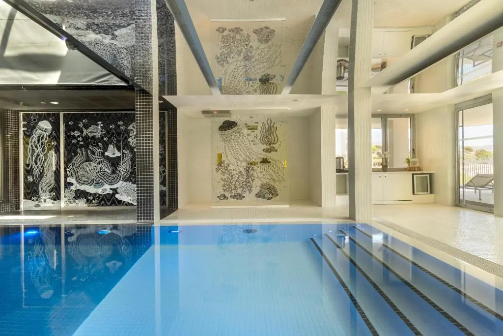 Piscine intérieure proposant une ambiance séminaire bien-être, comprenant un bassin rectangulaire bleu vif avec marches carrelées au premier plan, des fresques en mosaïque à motifs marins de méduses et pieuvres sur des murs carrelés noirs au centre, et un espace lumineux ouvert avec baies vitrées menant à une terrasse extérieure en arrière-plan.