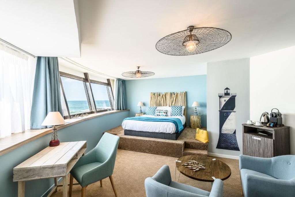 Chambre d'hôtel avec estrade de lit surélevée à tête de lit façon bois flotté et linge de lit bleu et blanc au centre, suspensions façon filet accrochées au-dessus, fauteuils bleus autour d'une table basse ronde en bois au premier plan, fresque murale de phare et coin café en bois à droite, et bureau en bois avec chaise près de fenêtres donnant sur la mer à gauche, une chambre adaptée à un séminaire en bord de mer.