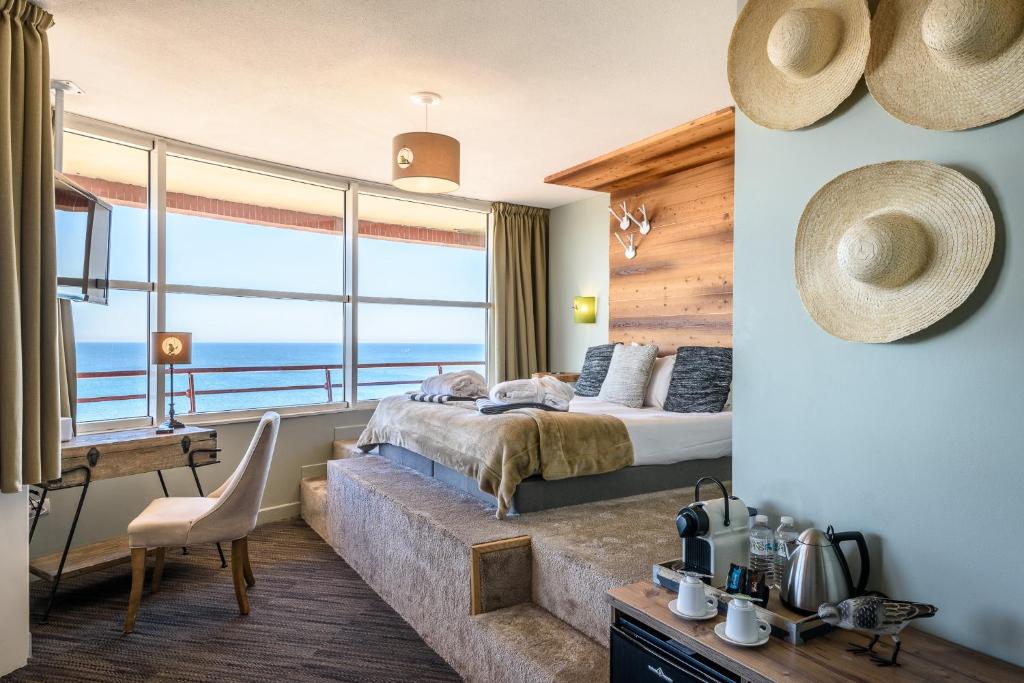 Chambre d'hôtel avec estrade de lit surélevée contre un mur en panneaux de bois formant tête de lit au centre, murs bleus décorés de trois chapeaux de paille à droite, coin café avec machine Nespresso et bouilloire au premier plan, bureau en bois et chaise rembourrée à gauche, et baies vitrées du sol au plafond donnant sur la mer en arrière-plan, une chambre adaptée à un séminaire en bord de mer.