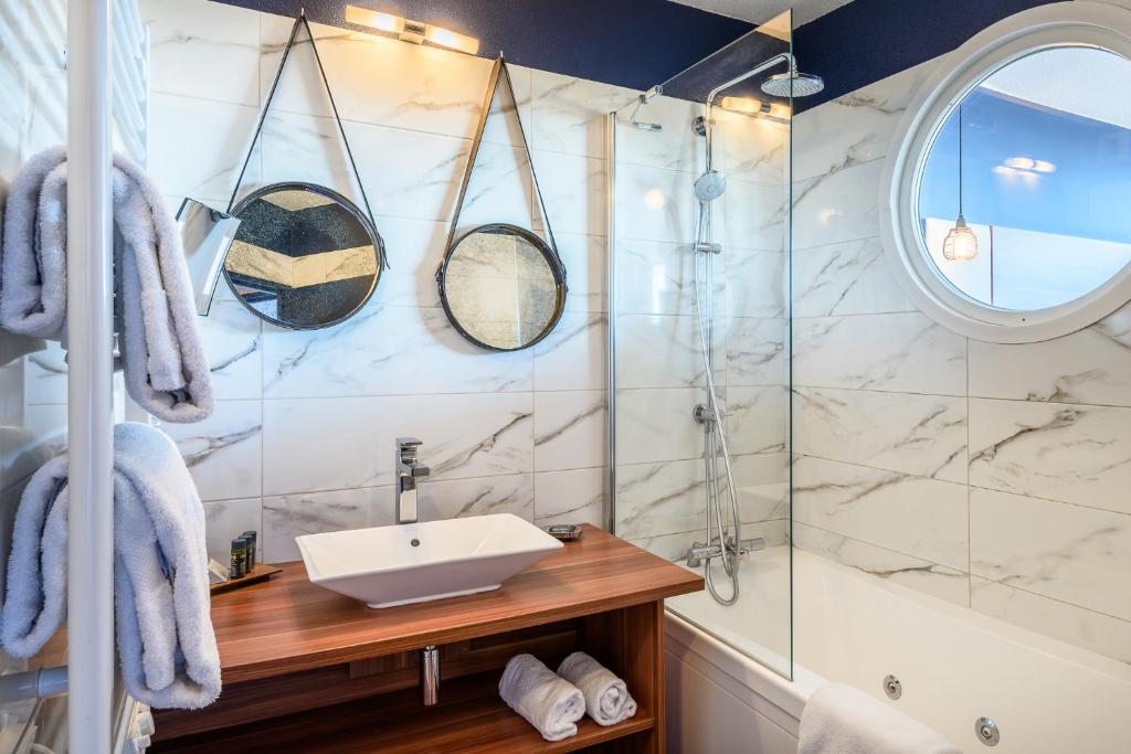 Salle de bain d'hôtel avec mur carrelé en marbre et deux miroirs ronds suspendus au-dessus d'un meuble-vasque en bois avec vasque blanche au premier plan, serviettes pliées sur un porte-serviettes et sur l'étagère en dessous, baignoire à paroi vitrée avec douche de tête à droite, et fenêtre ronde façon hublot tout à droite, adaptée à un séminaire en bord de mer.