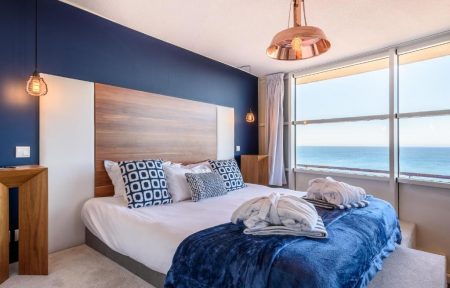 Chambre d'hôtel en bord de mer avec un lit habillé de linge blanc, des coussins bleu marine à motifs, des peignoirs blancs pliés et un jeté bleu marine au premier plan, une tête de lit en bois contre un mur bleu foncé avec suspensions en cuivre à gauche, et de grandes baies vitrées offrant une vue sur la mer en arrière-plan.