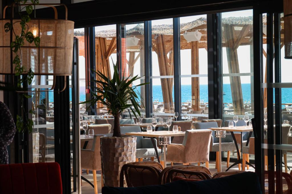 Restaurant sur place d'hôtel avec des tables en bois dressées de verres à vin et des chaises rembourrées beige au centre, une grande plante verte en pot dans un cache-pot effet pierre au premier plan, une suspension en fibres tressées avec lierre retombant à gauche, et des baies vitrées du sol au plafond révélant une terrasse à pergola en bois et une vue sur la mer en arrière-plan.