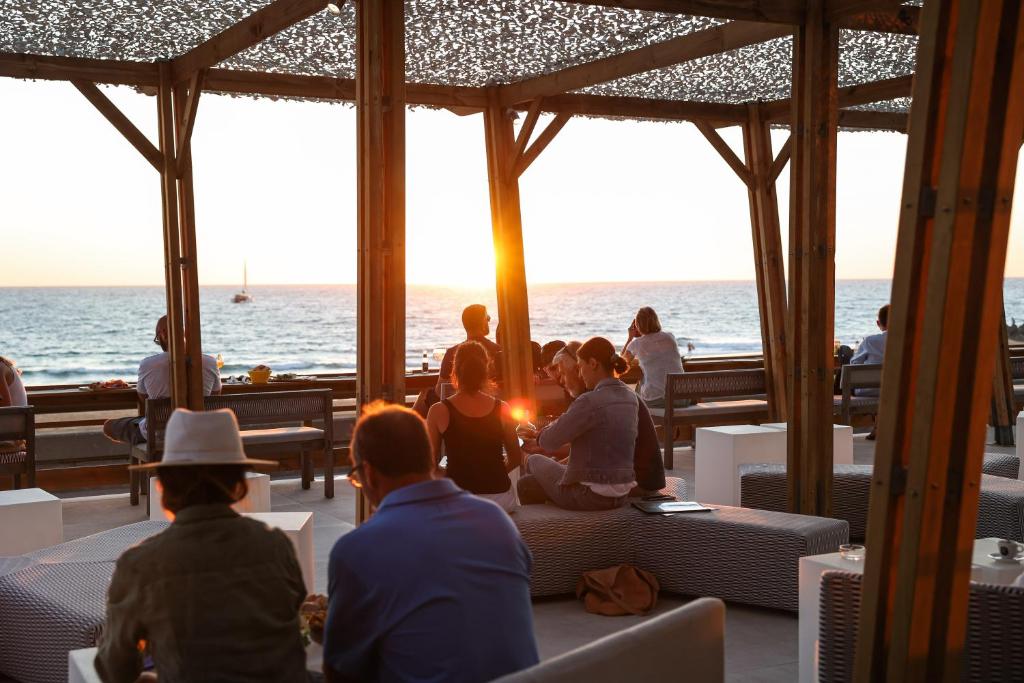 Terrasse afterwork en bord de mer avec des personnes assises sur des bancs tressés gris et des tables cubiques blanches au premier plan, des groupes discutant sous une structure en pergola en bois au centre, et la mer avec un voilier visible sous un soleil couchant en arrière-plan.