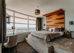 Chambre d'hôtel séminaire en bord de mer avec un lit habillé de linge gris et des coussins à motifs au premier plan, une estrade surélevée avec une tête de lit en bois ornée de crochets en forme de bois de cerf et des appliques murales vertes au centre, un fauteuil rembourré et un petit bureau avec lampe à gauche, et de grandes fenêtres avec vue sur la mer en arrière-plan.
