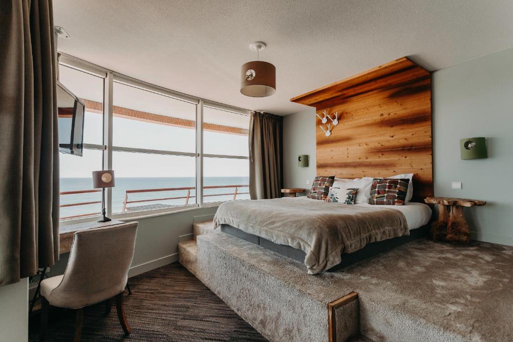 Chambre d'hôtel séminaire en bord de mer avec un lit habillé de linge gris et des coussins à motifs au premier plan, une estrade surélevée avec une tête de lit en bois ornée de crochets en forme de bois de cerf et des appliques murales vertes au centre, un fauteuil rembourré et un petit bureau avec lampe à gauche, et de grandes fenêtres avec vue sur la mer en arrière-plan.