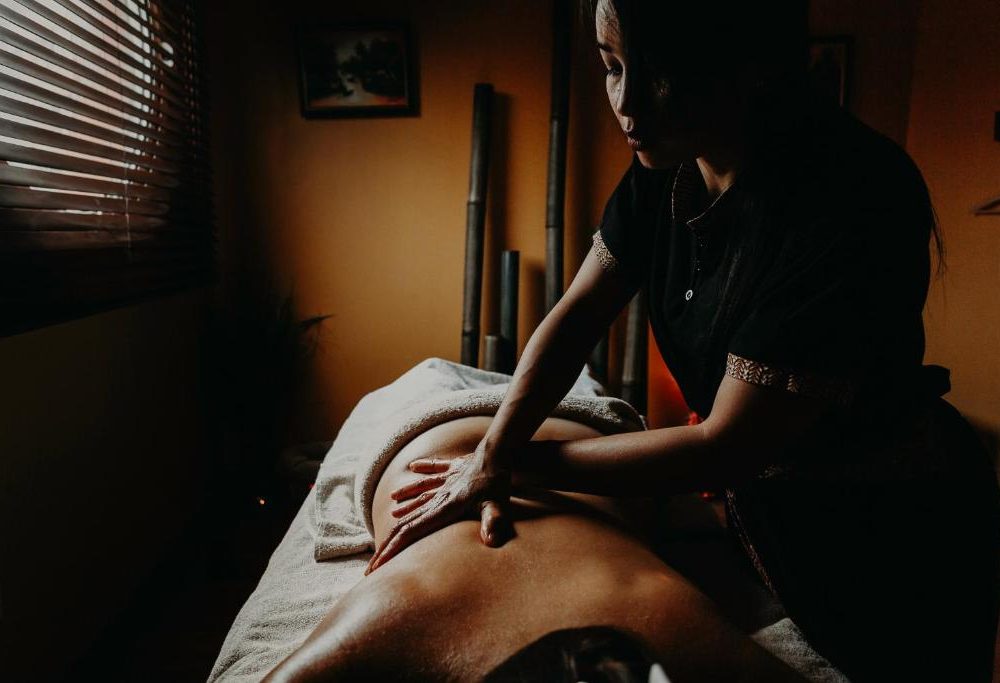 Gros plan sur un soin spa de séminaire bien-être avec une personne recevant un massage du dos sur une table à serviettes grises au premier plan, une praticienne en tenue sombre effectuant le massage au centre, des bâtons de bambou décoratifs et un cadre photo contre un mur éclairé en orange à gauche, et des stores en bois à lattes en arrière-plan sous un éclairage tamisé et chaleureux.