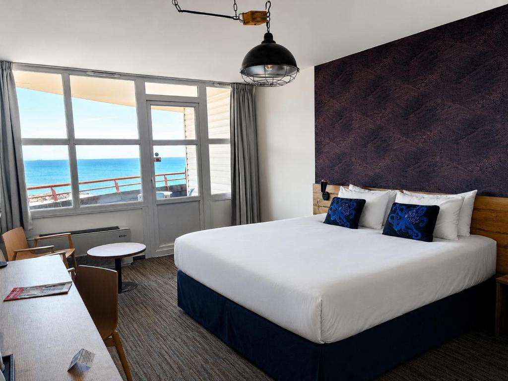 Chambre d'hôtel avec lit double aux draps blancs et soubassement bleu marine et coussins à motif de pieuvre contre un mur d'accent sombre texturé à droite, tête de lit en bois et suspension style industriel au-dessus, bureau en bois et chaises à gauche, et portes-fenêtres et fenêtres donnant sur la mer et un balcon en arrière-plan, une chambre adaptée à un séminaire en bord de mer.
