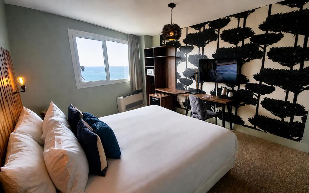 Chambre d'hôtel en bord de mer avec un lit habillé de linge blanc et des coussins bleu marine et beige au premier plan, une tête de lit en lattes de bois avec applique murale à gauche, un bureau en bois avec chaise, bouilloire et télévision murale à droite sur un papier peint à motif d'arbres noir et blanc, et une fenêtre avec vue sur la mer et rideaux en arrière-plan.